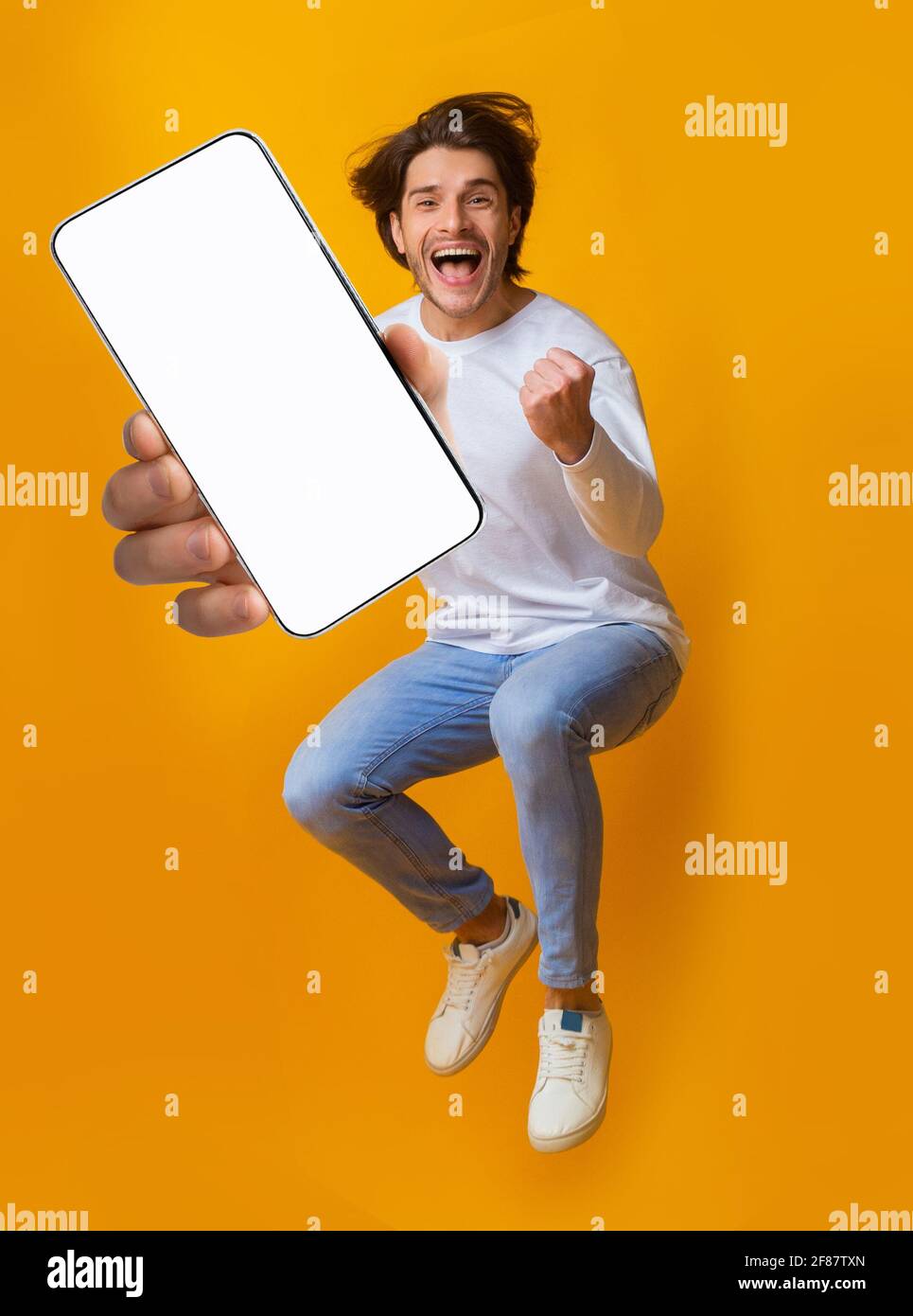 Neue tolle mobile App. Springen emotionaler junger Mann zeigt Smartphone mit leerem Bildschirm auf orangefarbenem Hintergrund, Mockup Stockfoto