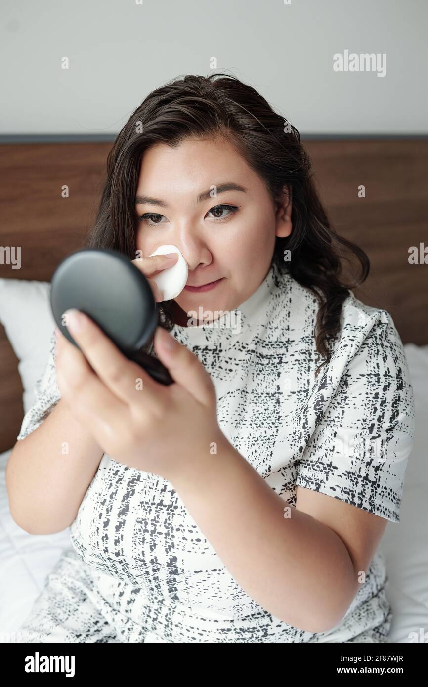 Schöne junge Frau, die auf dem Bett sitzt und ein durchscheinendes Gesicht aufführt Puder beim Make-up Stockfoto