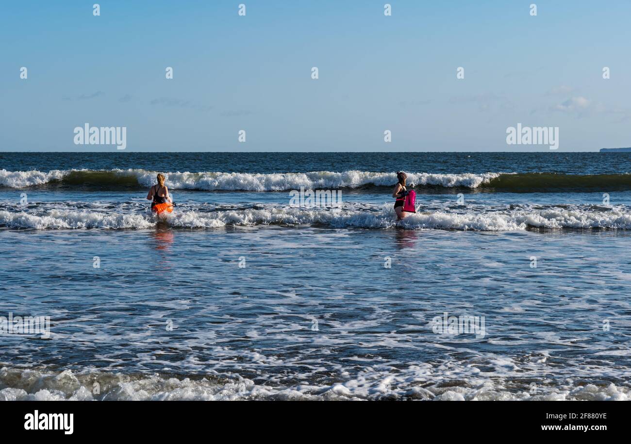 Zwei Schwimmerinnen im Freien oder im Freien in Badeanzügen mit Schwimmern gehen an einem sonnigen Tag ins Meer, Firth of Forth, Schottland, Großbritannien Stockfoto