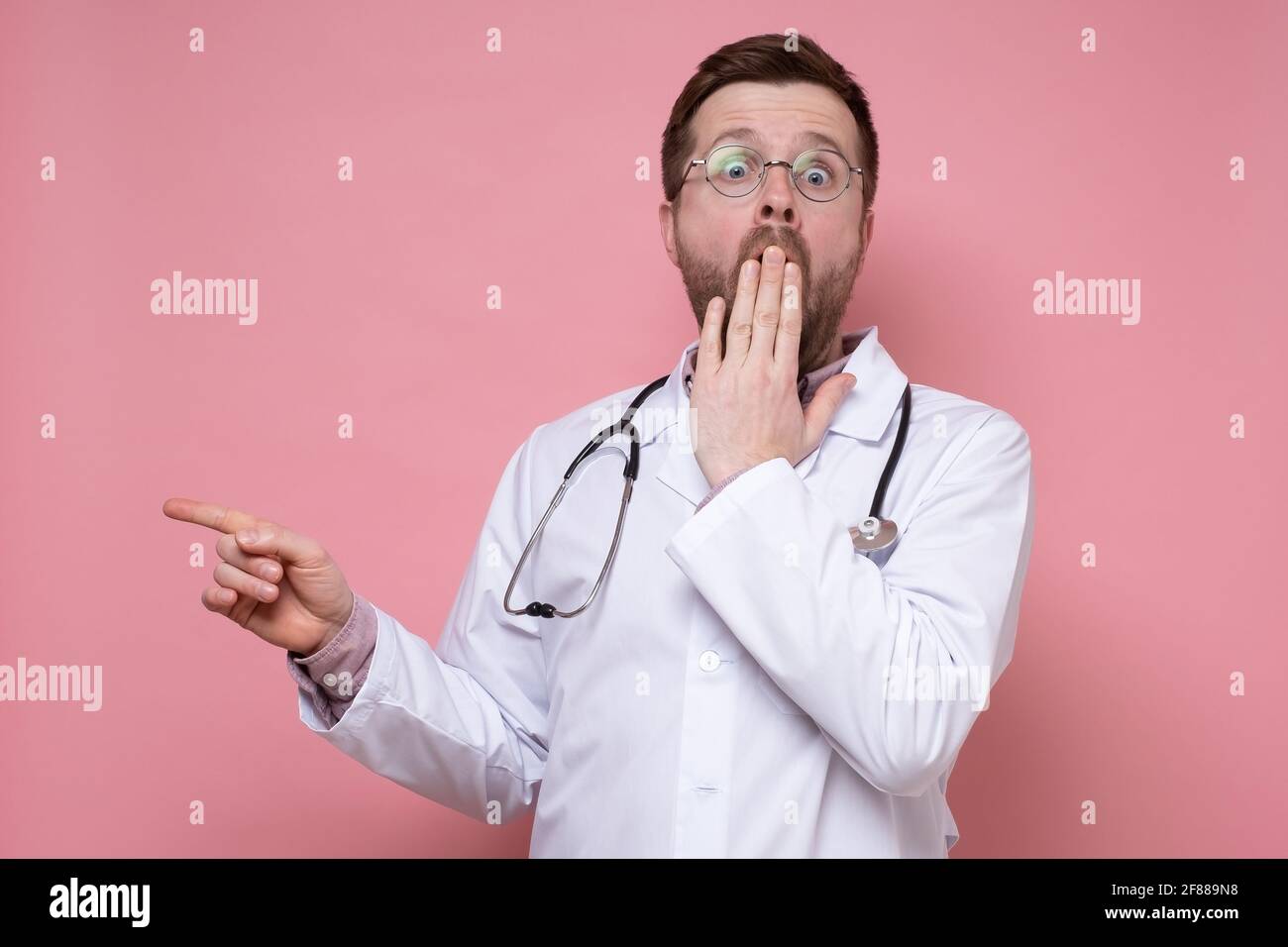 Geschockter Arzt mit einem Stethoskop um den Hals, erschrocken, zeigt den Finger zur Seite, blickt auf die Kamera und bedeckt den Mund mit der Hand. Stockfoto
