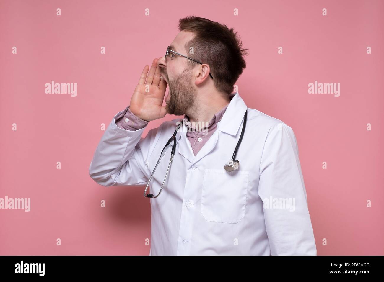 Ein junger Arzt mit einem Stethoskop um den Hals ruft jemanden an, er schreit laut von Hand zu Mund und schaut weg. Rosa Hintergrund. Stockfoto