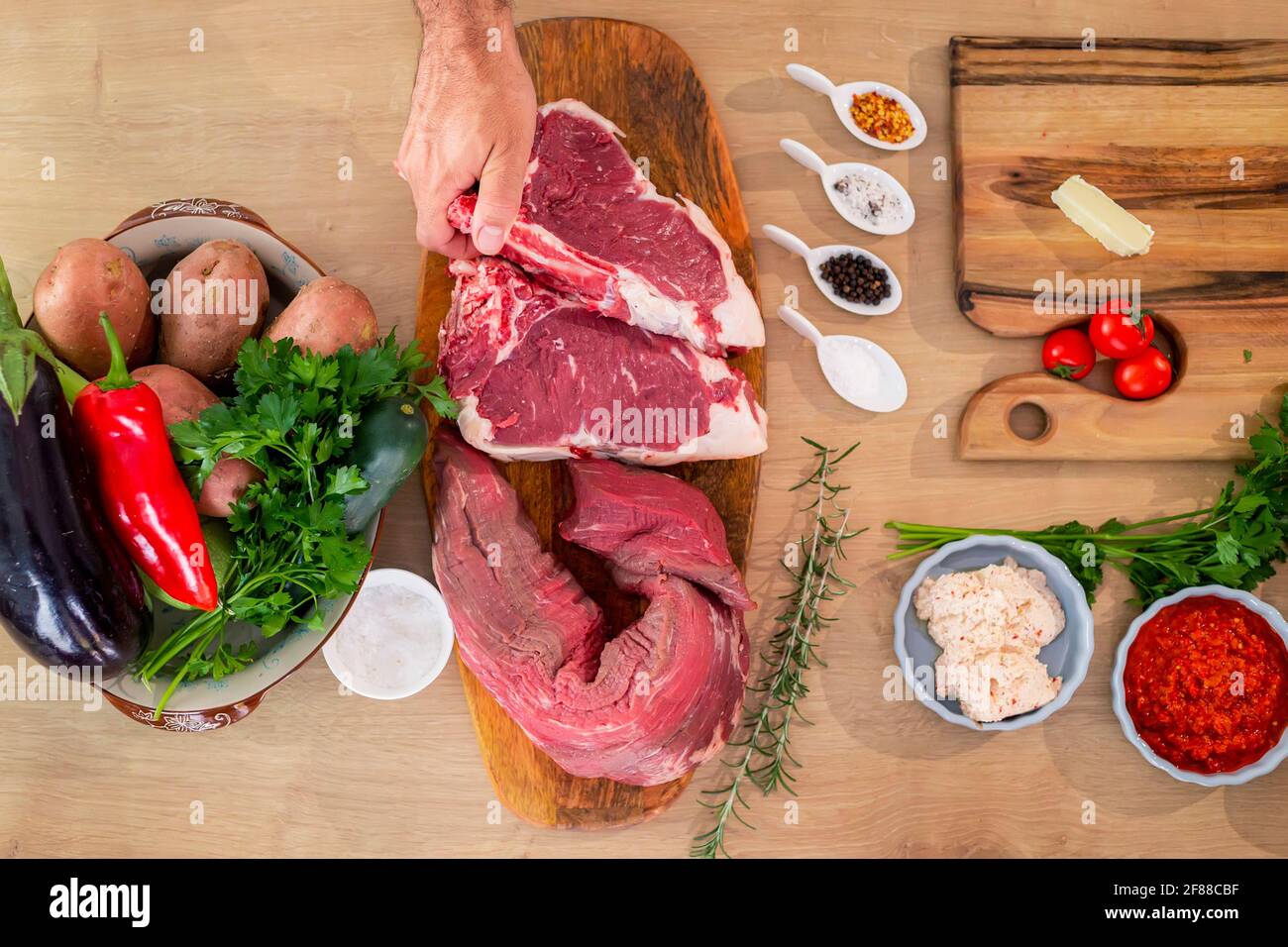 Fleischstücke, Gewürze und Gemüse von oben auf einem hölzernen Küchentafel fotografiert. Auf der Seite stehen Kartoffeln, Paprika und Kürbisse in einem Topf Stockfoto