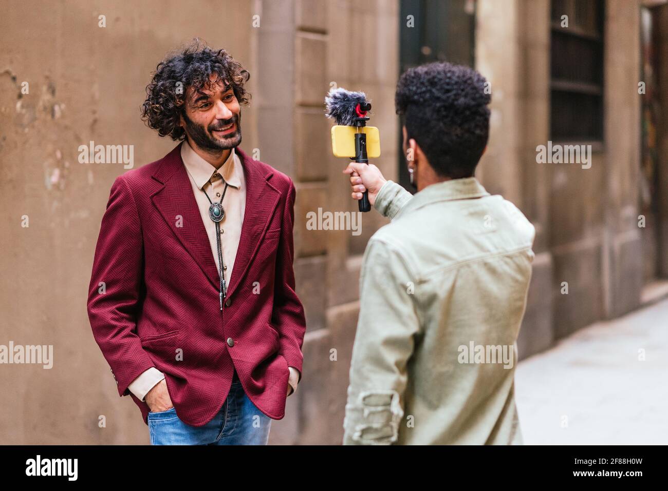 Ein junger, trendiger Mann aus Kaukasien, der auf der Straße bei einer Live-Veranstaltung steht. Er wird von einem lateinischen Kollegen mit einem Smartphone aufgezeichnet, das an einem Stabilisator befestigt ist. Stockfoto