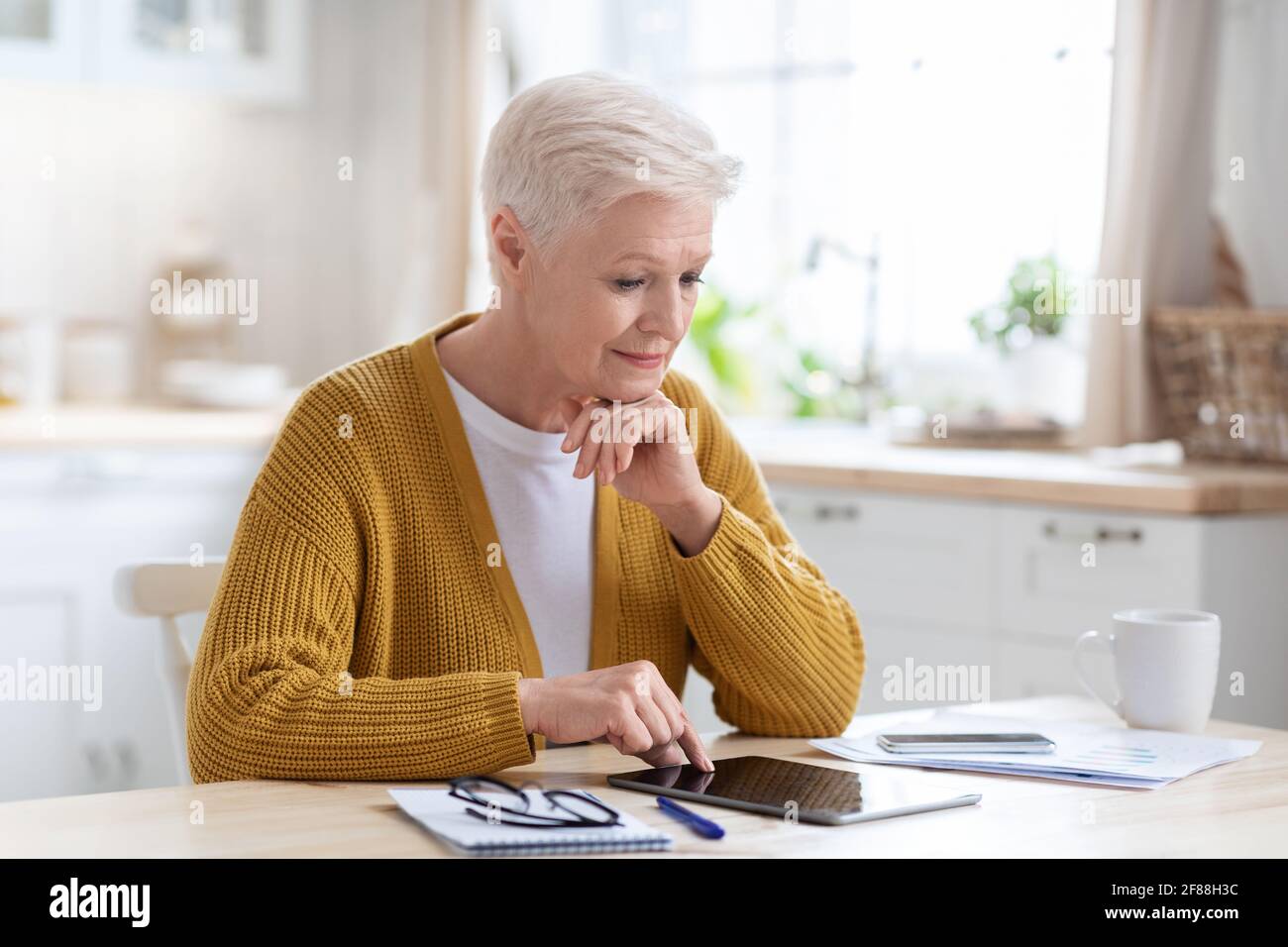 Ältere Geschäftsfrau, die in der Küche sitzt und ein Tablet benutzt Stockfoto Ältere Geschäftsfrau, die in der Küche sitzt und ein Tablet benutzt Stockfoto