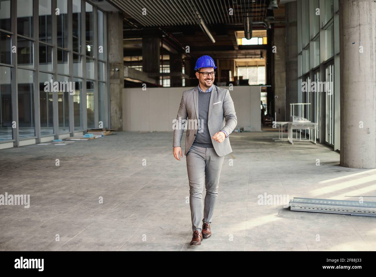 Lächelnder Geschäftsmann mit Helm auf dem Kopf, der durch die Baustelle seines Gebäudes läuft. Stockfoto