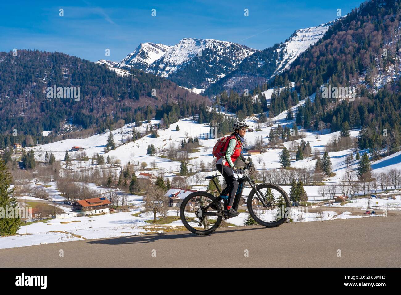 Senior Frau Mountainbiken unter der Nagelfluh-Bergkette mit Hochgrat-Gipfel auf einem E-Mountainbike im Frühfrühling, im Allgäu bei Stei Stockfoto