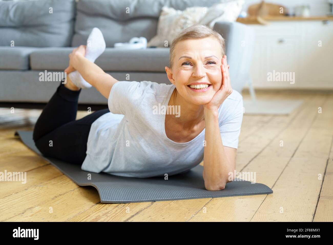 Glückliche lächelnde ältere Frau, die sich streckte und zur Kamera schaute. Bewegung für die körperliche Gesundheit. Wellness für Frauen im Ruhestand. Inländische Yoga-Praxis der flexiblen reifen Frau Ausübung auf Sportmatte Stockfoto