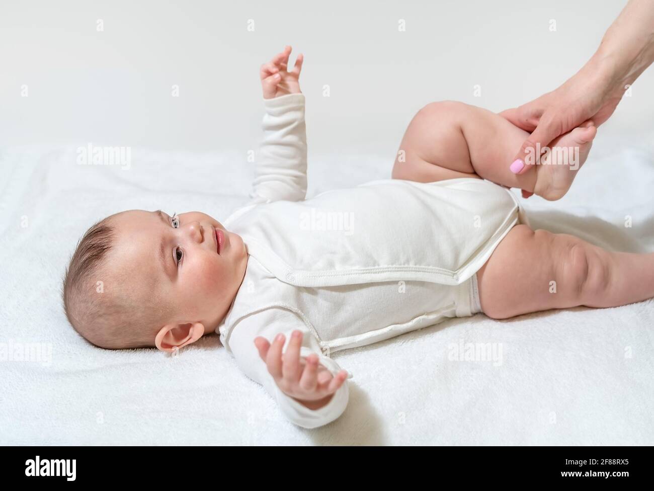 Neugeborenes gesundes Baby, das körperliche Übungen für die Beine und das Hüftgelenk macht. Mit Hilfe der Mutter des Sanitäters. Auf weißem Hintergrund. Stockfoto