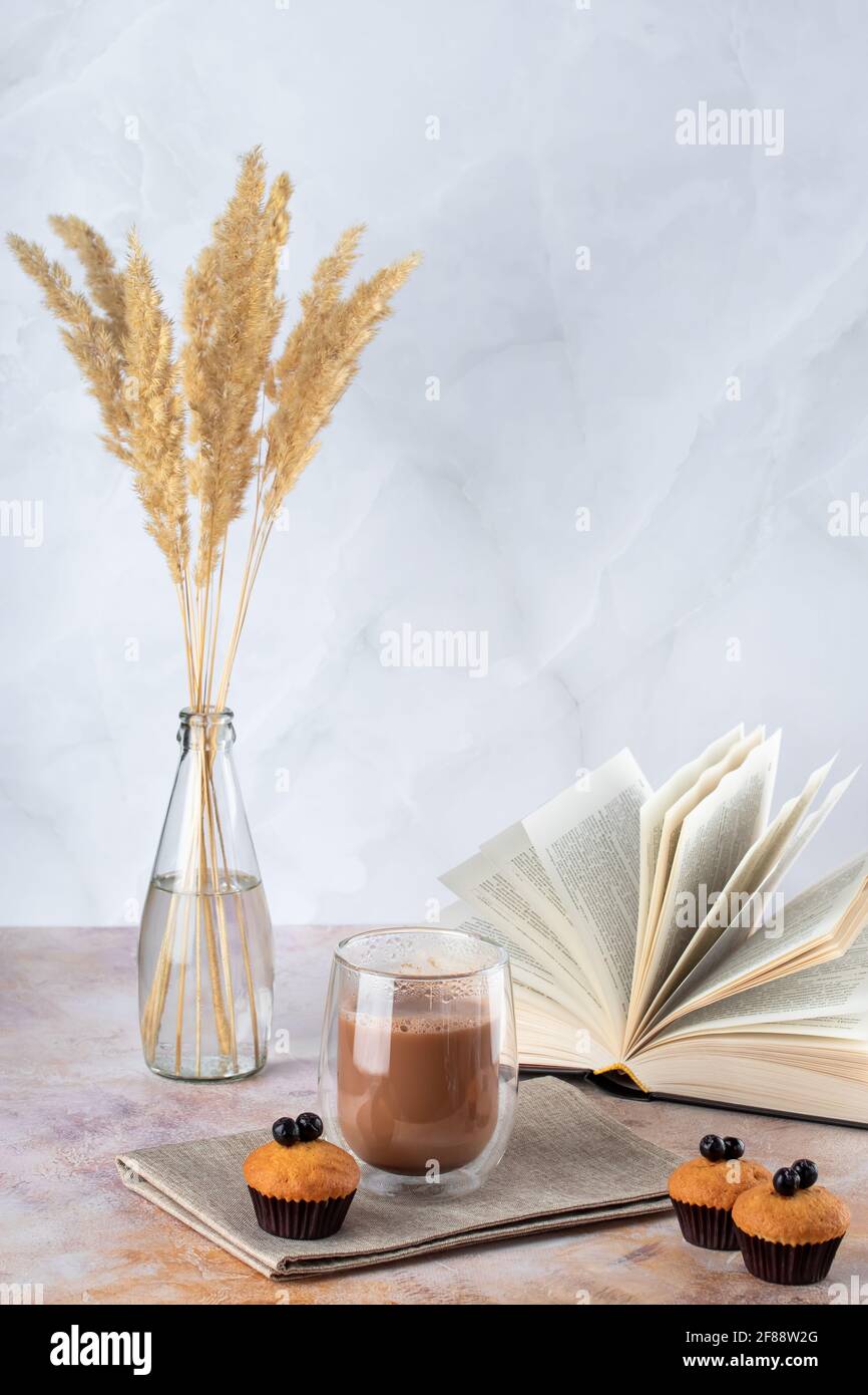 Muffins und ein Glas Kaffee mit Milch auf dem Tisch. Heißes Getränk, Kakao und offenes Buch. Getrocknetes Pampagras in Glasflasche als Vase. Natürliche Pflanzen Dekor o Stockfoto