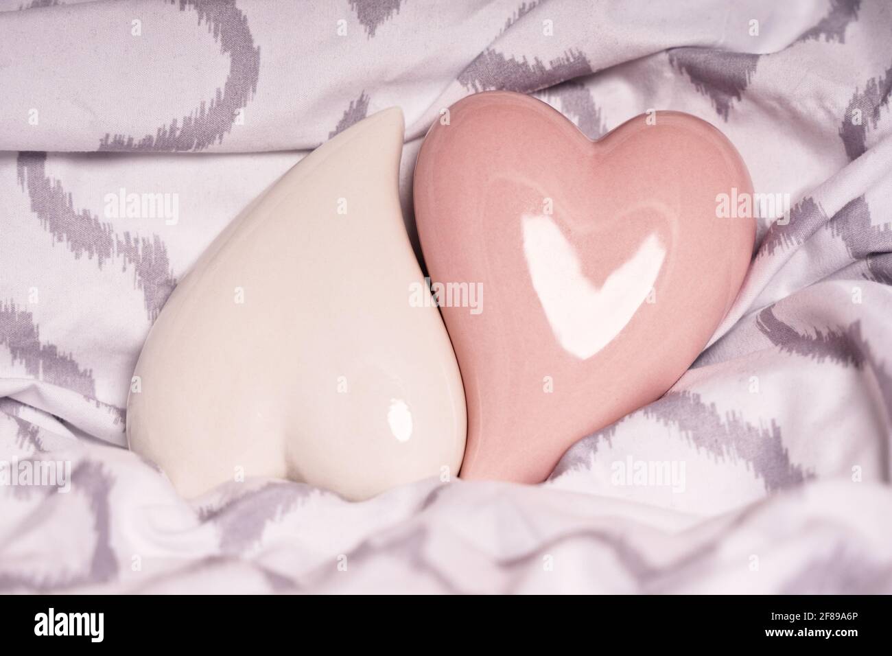 Herzförmige Keramikdekorationen, die auf einem modernen Kissen oder Bettlaken ineinander passen. Zwei rote Herzen auf einem Bett. Valentinstag, Hochzeitskonzept Stockfoto