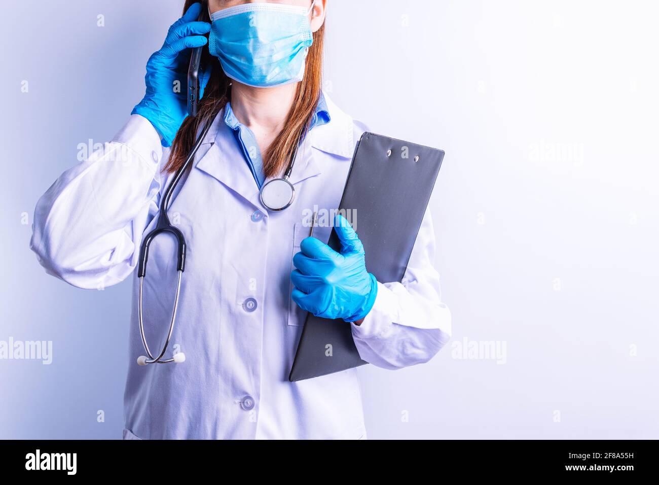 Kaukasische junge Frau trägt Stethoskop Kleid und Handschuhe und Maske hält Handy an einem Ohr und Berichte im anderen. Technologie und Medizin c Stockfoto