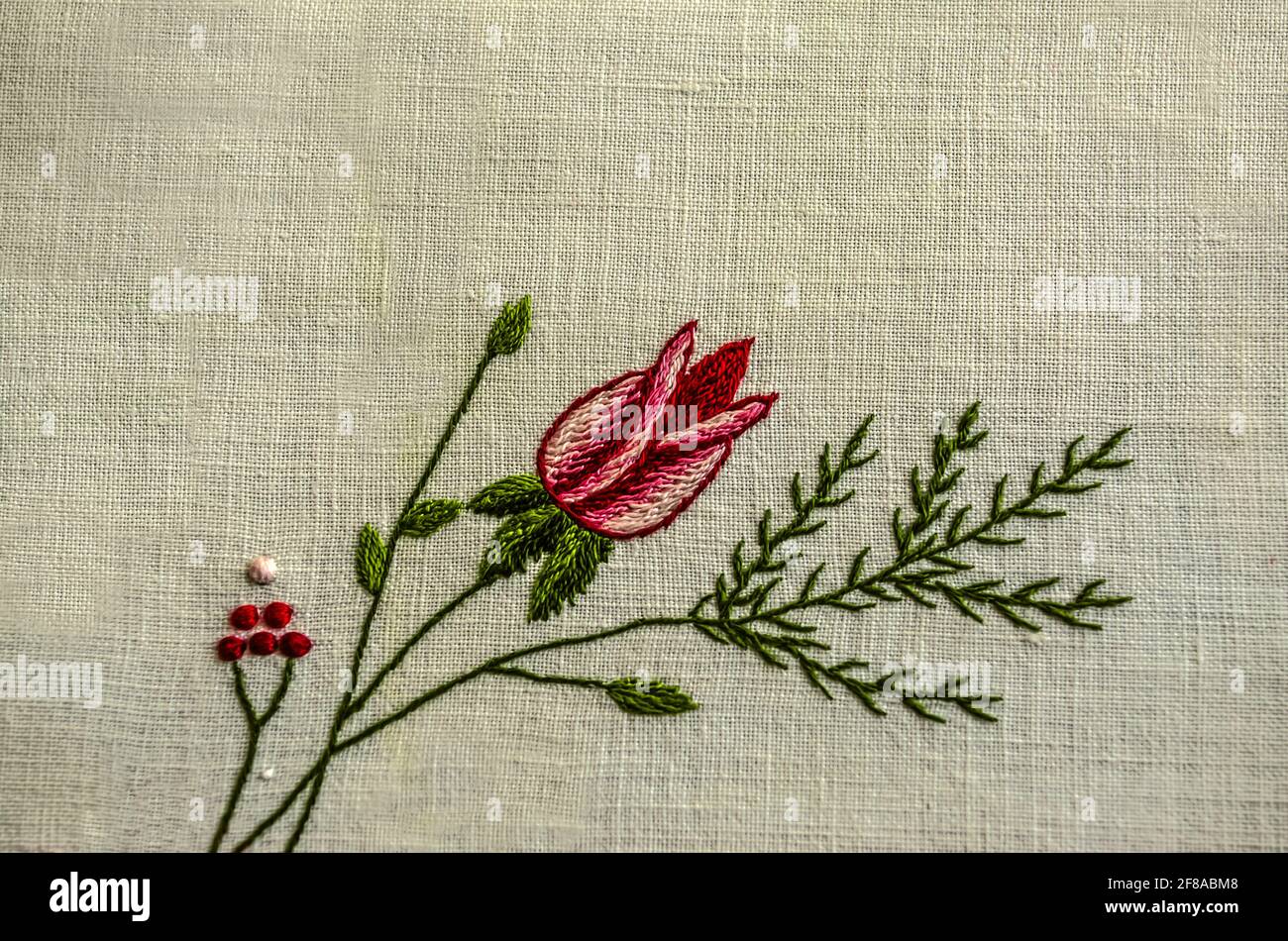 Bestickter Zweig mit einer roten Rosenknospe, einem Nadelzweig und kleinen roten Beeren auf einem weißen Baumwollstoff. Stockfoto