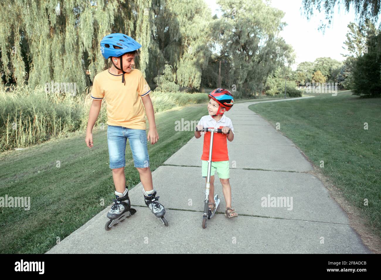 Kaukasische Jungen Brüder in Helmen Reiten Rollschuhe und Roller auf der Straße im Park. Saisonale Outdoor-Aktivitäten für Kinder Sport. Gesundes Kindheitsleben Stockfoto