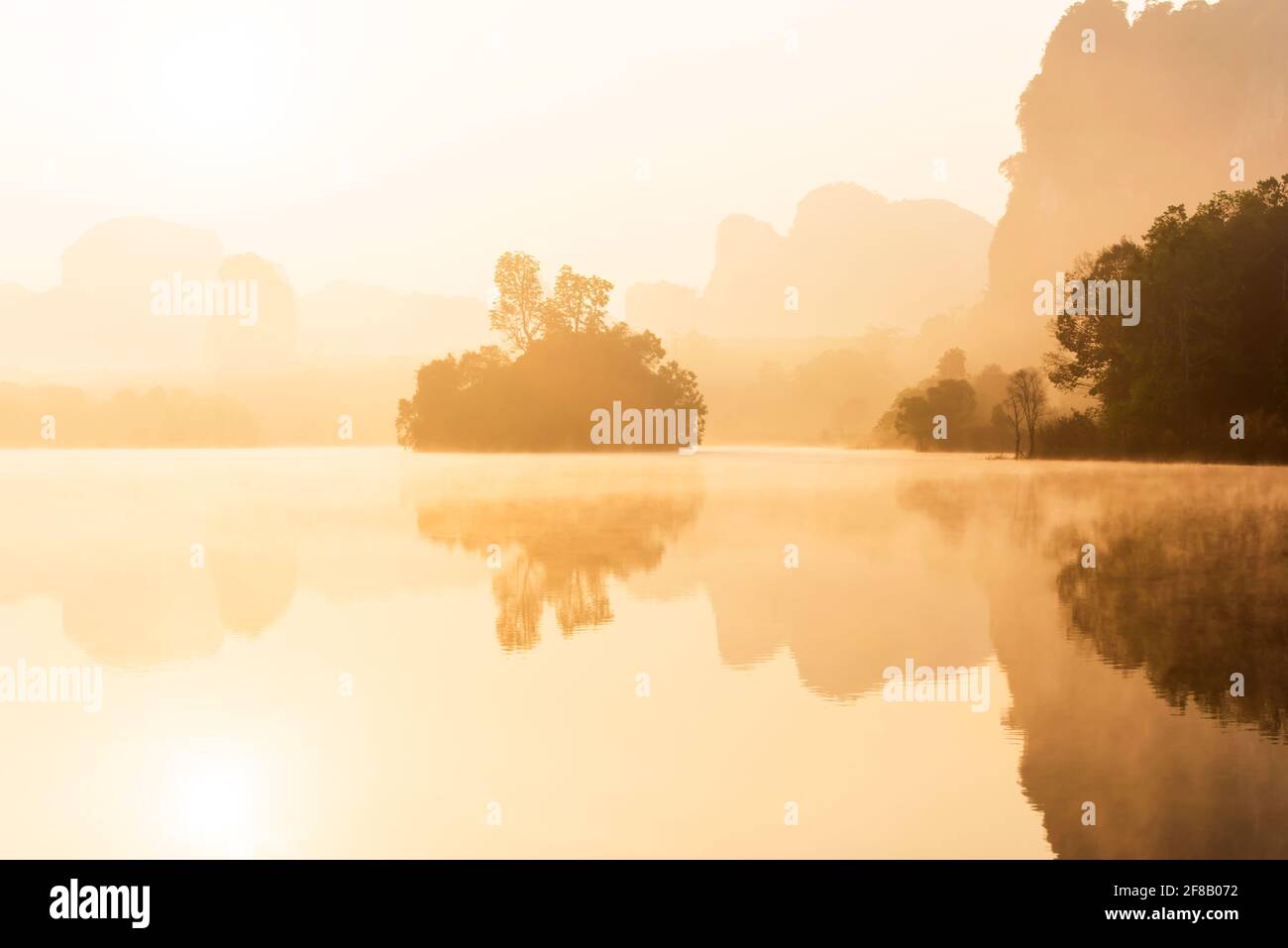 Landschaftslandschaft mit tropischem See im Morgennebel, goldener Sonne, die auf dem See und der Bergkette im Hintergrund scheint. Long Talay, Thailand. Stockfoto