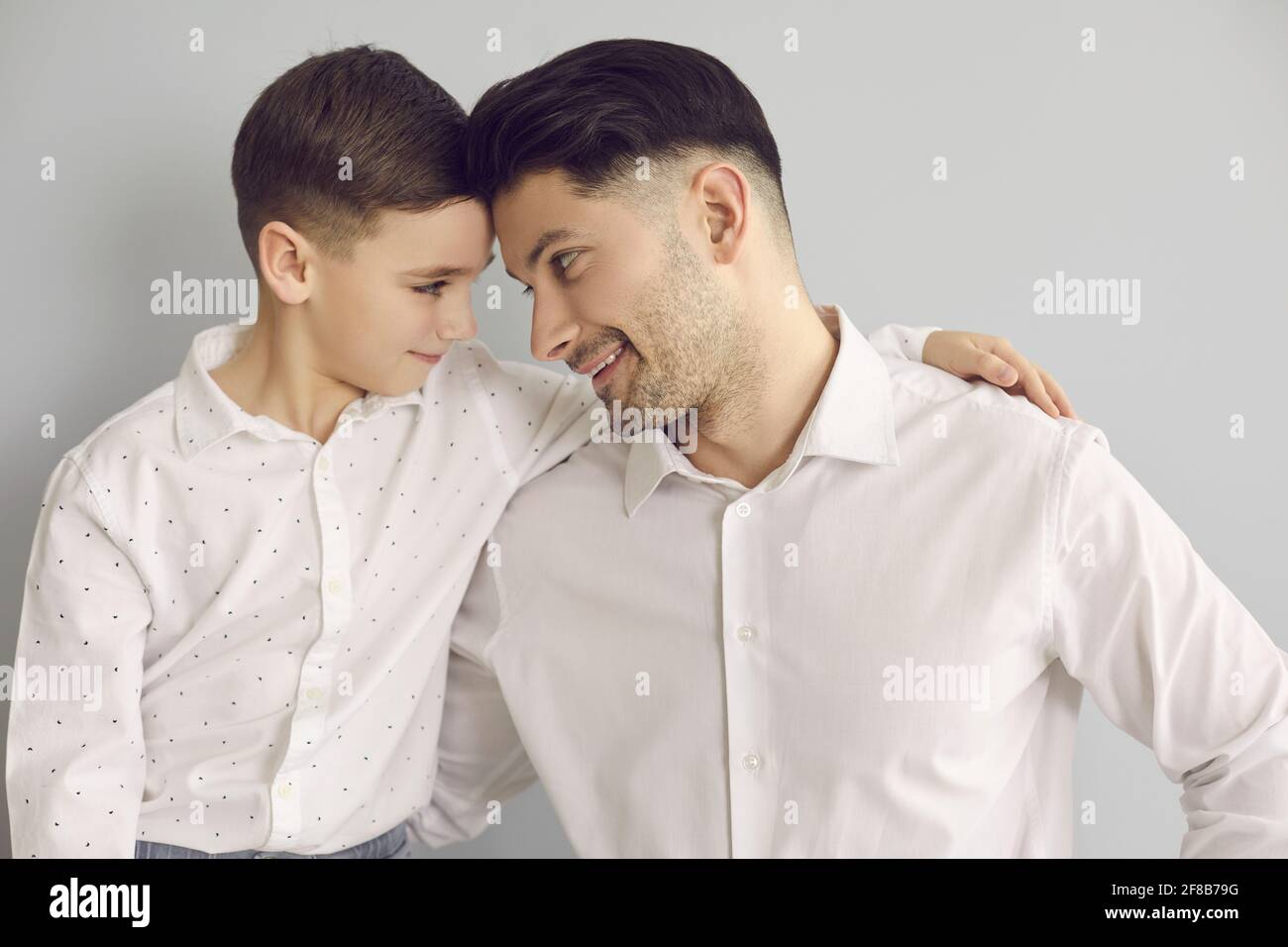 Porträt eines Vaters mit einem kleinen Sohn, der sich umarmt und liebevoll auf grauem Hintergrund anschaut. Stockfoto