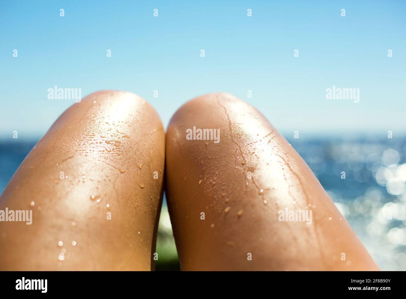Glatte, gebräunte weibliche Beine im Wasserstrahl am Strand vor dem Hintergrund des Meeres. Bräunungsprodukte, Sonnenbrandschutz, Hautpflege, Pflegemittel Stockfoto