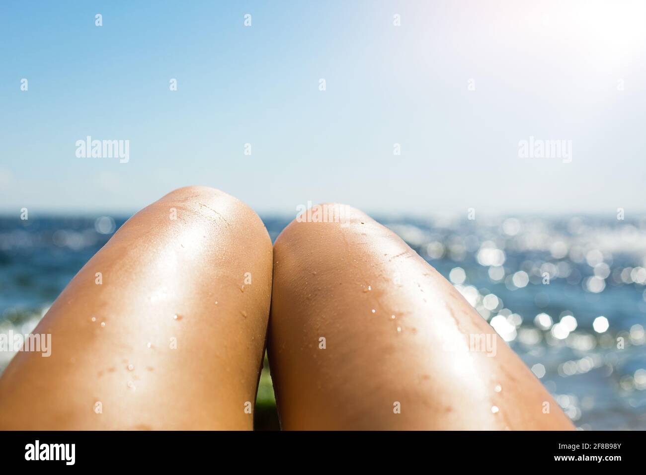Glatte, gebräunte weibliche Beine im Wasserstrahl am Strand vor dem Hintergrund des Meeres. Bräunungsprodukte, Sonnenbrandschutz, Hautpflege, Pflegemittel Stockfoto