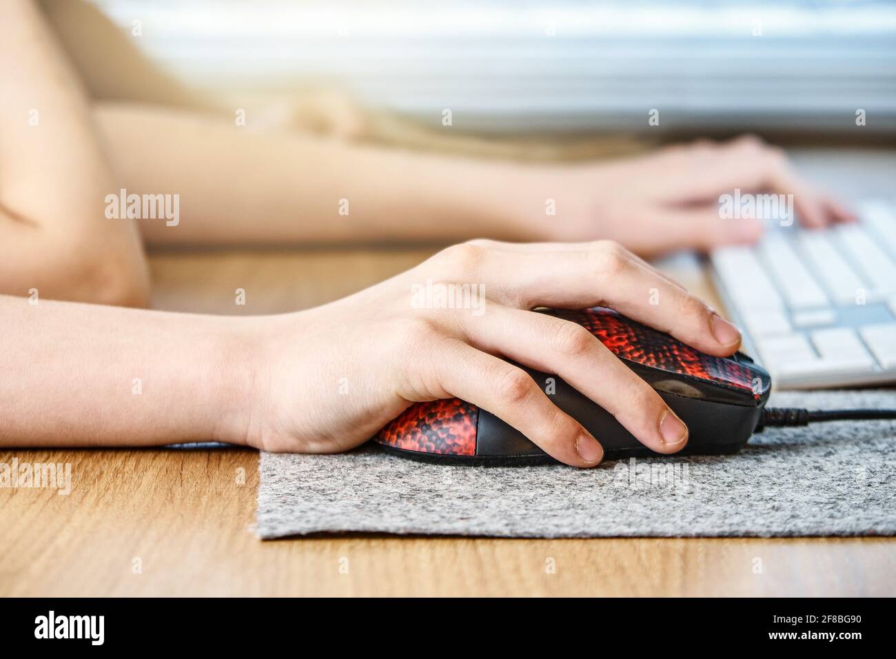 Junge Schuljunge Hände auf rote schwarze Maus mit Filzpad Und verschwommenes Keyboard in weißer Farbe auf braunem Holztisch Nahaufnahme Stockfoto