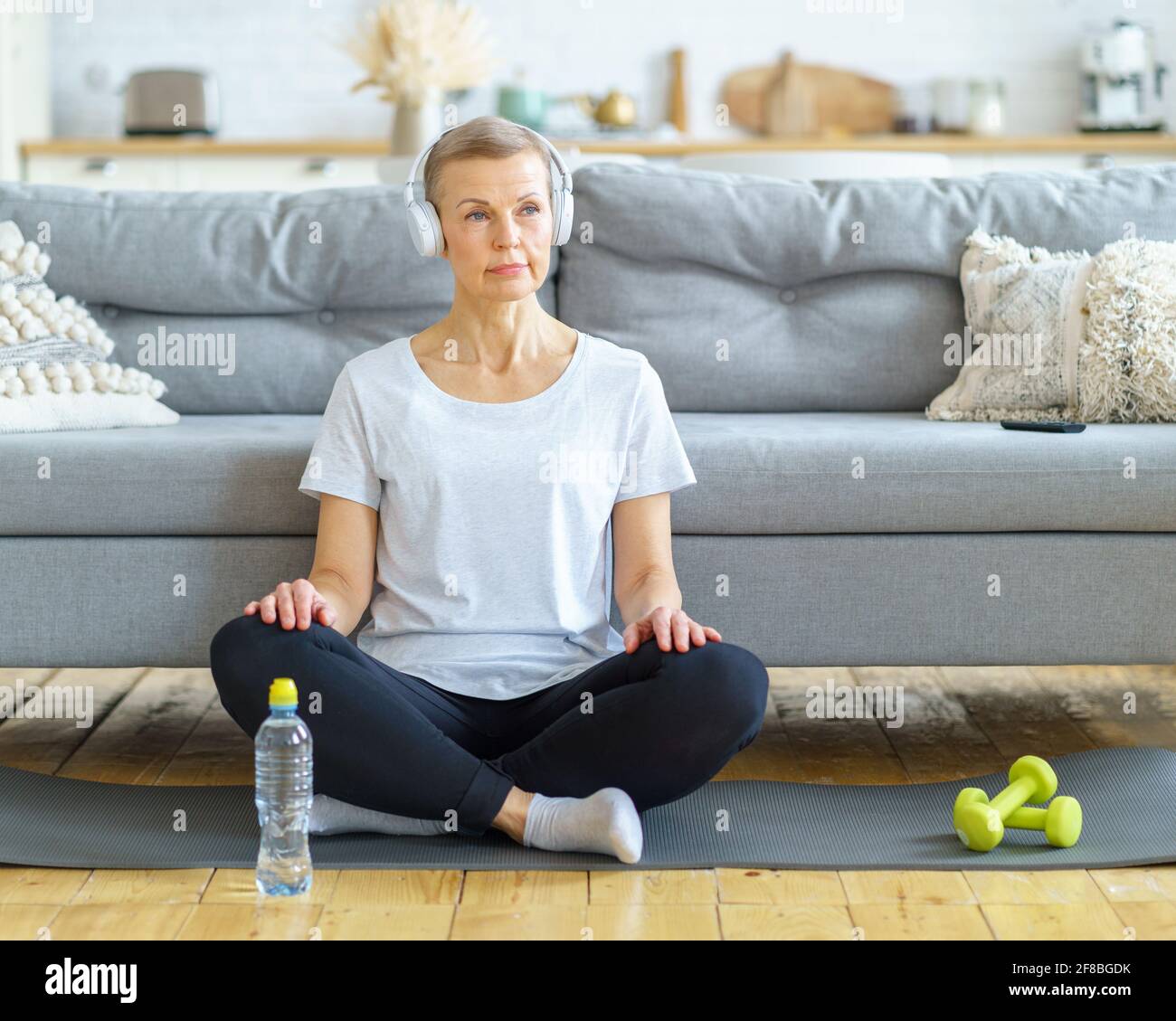 Ältere Frau sitzt mit gekreuzten Beinen in Kopfhörern und hört im Wohnzimmer Musik. Entspannen Sie sich nach dem Fitnesstraining. Emotionale und spirituelle Gesundheit. Wohlbefinden, Wellness mit Meditation. Genießen Sie schöne Momente Stockfoto