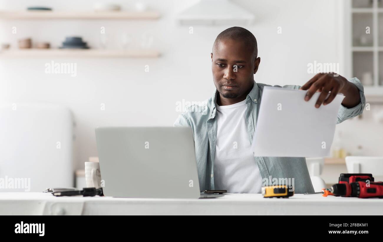 Ernsthafter schwarzer Handwerker am Schreibtisch mit Laptop und Werkzeug für Reparatur lesen Spezifikation und Vergleich mit Online-Handbuch Stockfoto