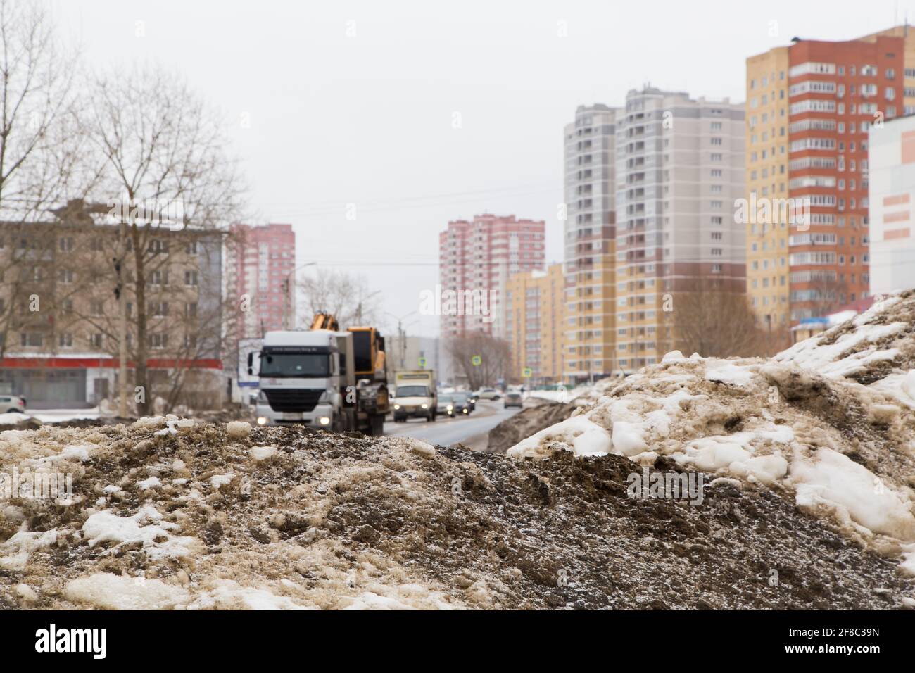 Große Schneeverwehung am Straßenrand mit vorbeifahrenden Autos. Auf einer Stadtstraße liegt ein Schneeblock mit schmutzigen Klumpen vor dem Hintergrund von Hochhäusern und einem grauen Himmel. Stockfoto