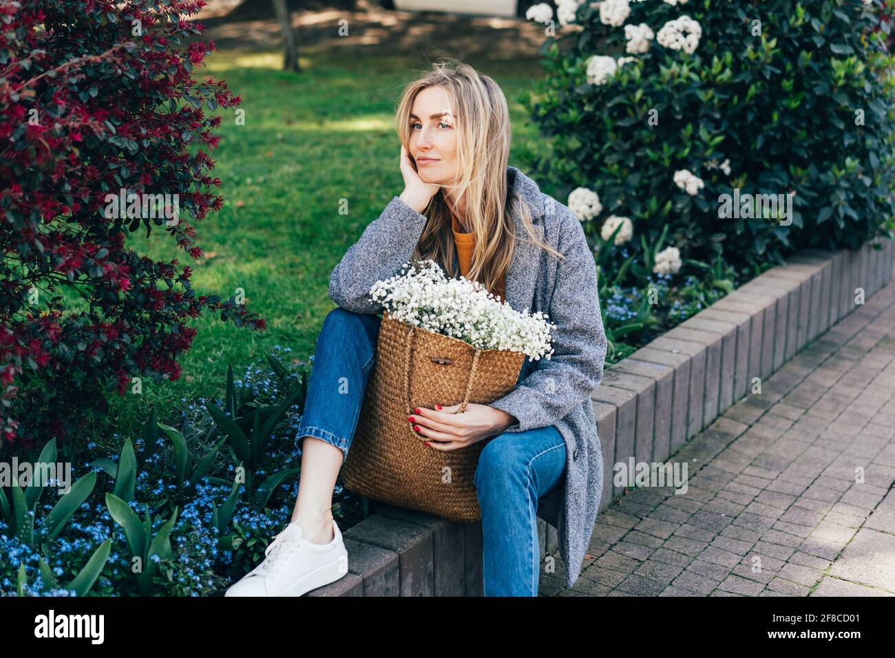 Ziemlich gelangweilte Frau in grauem Mantel, die neben der Blume sitzt Bett und Träumen..stilvolle kaukasische Frau posiert im Freien unter den Blumen Und trägt einen schönen Stockfoto