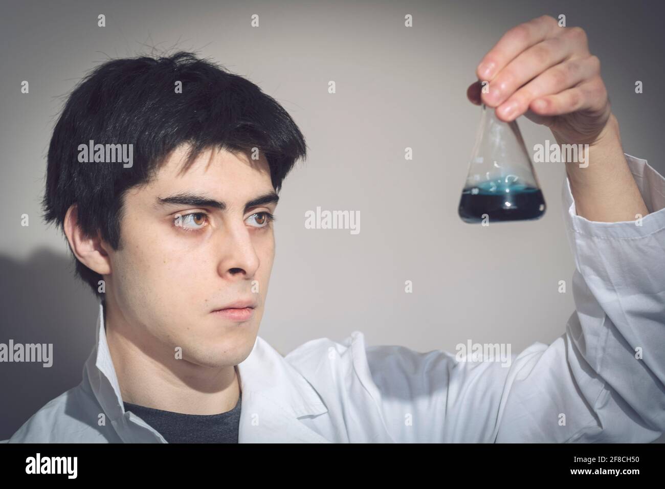 Ein junger kaukasischer Mann mit hispanischer Erscheinung in einem Labormantel, der eine Probe in einem konischen Kolben untersucht, Gymnasium, Universität, Chemie Stockfoto