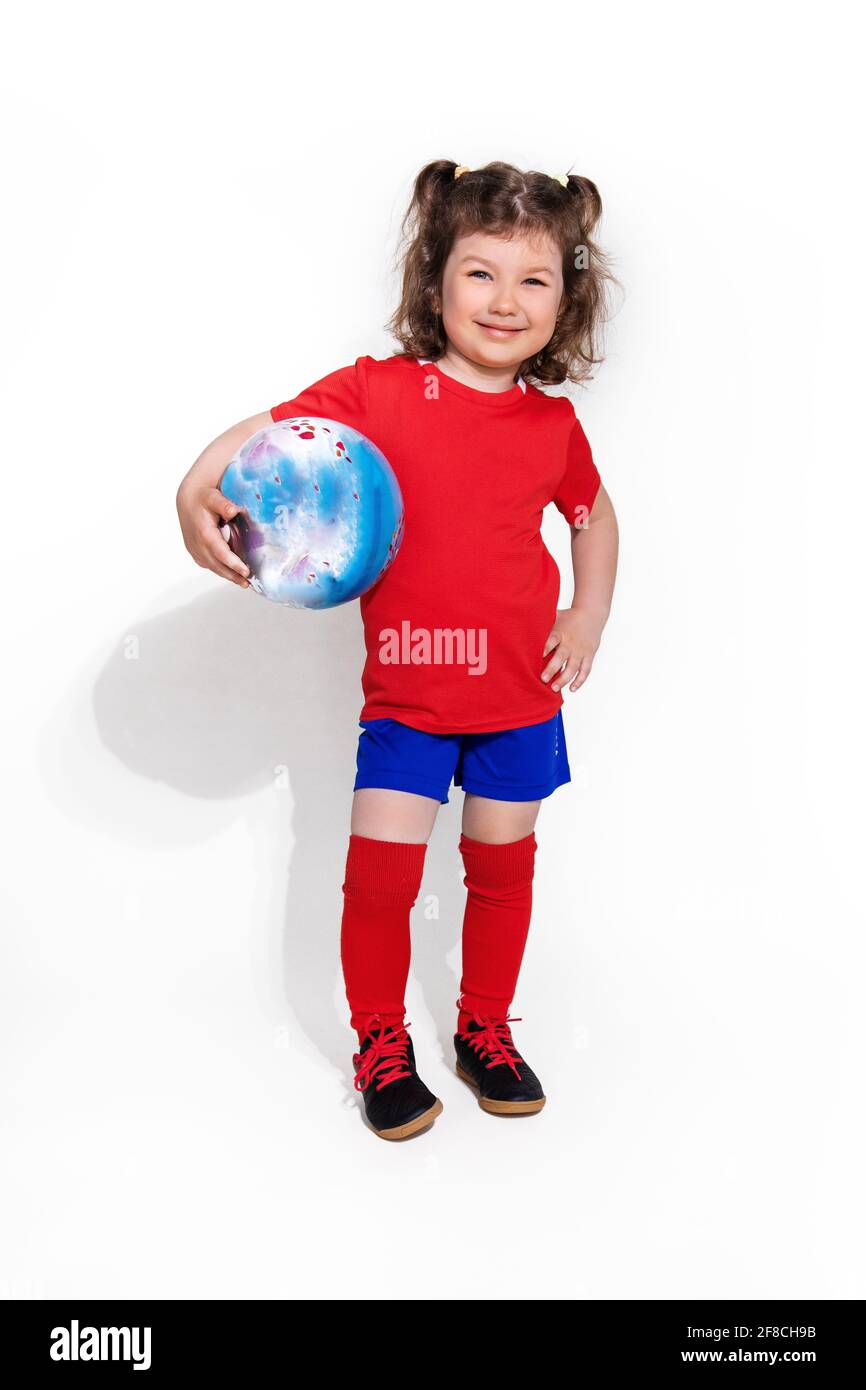 Kleines schönes Mädchen in Fußballuniform und mit Ball in der Hand auf weißem Hintergrund schaut in die Kamera. Frauenfußballkonzept für Kinder Stockfoto