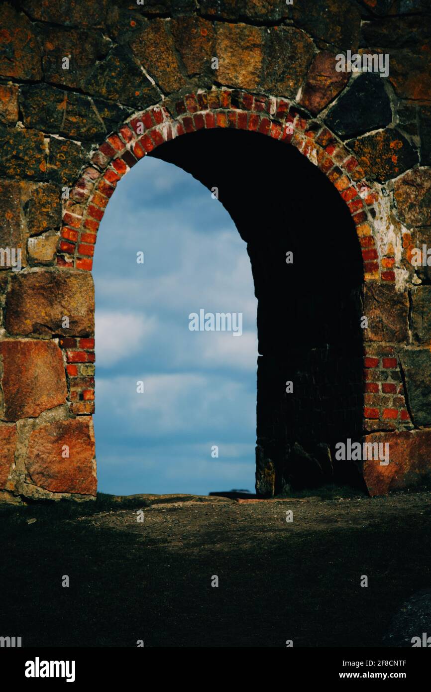 Ein alter Steinbogen öffnet sich wie ein Durchgang in die andere Welt, da man auf der anderen Seite den blauen, wolkigen Himmel sehen kann. Stockfoto