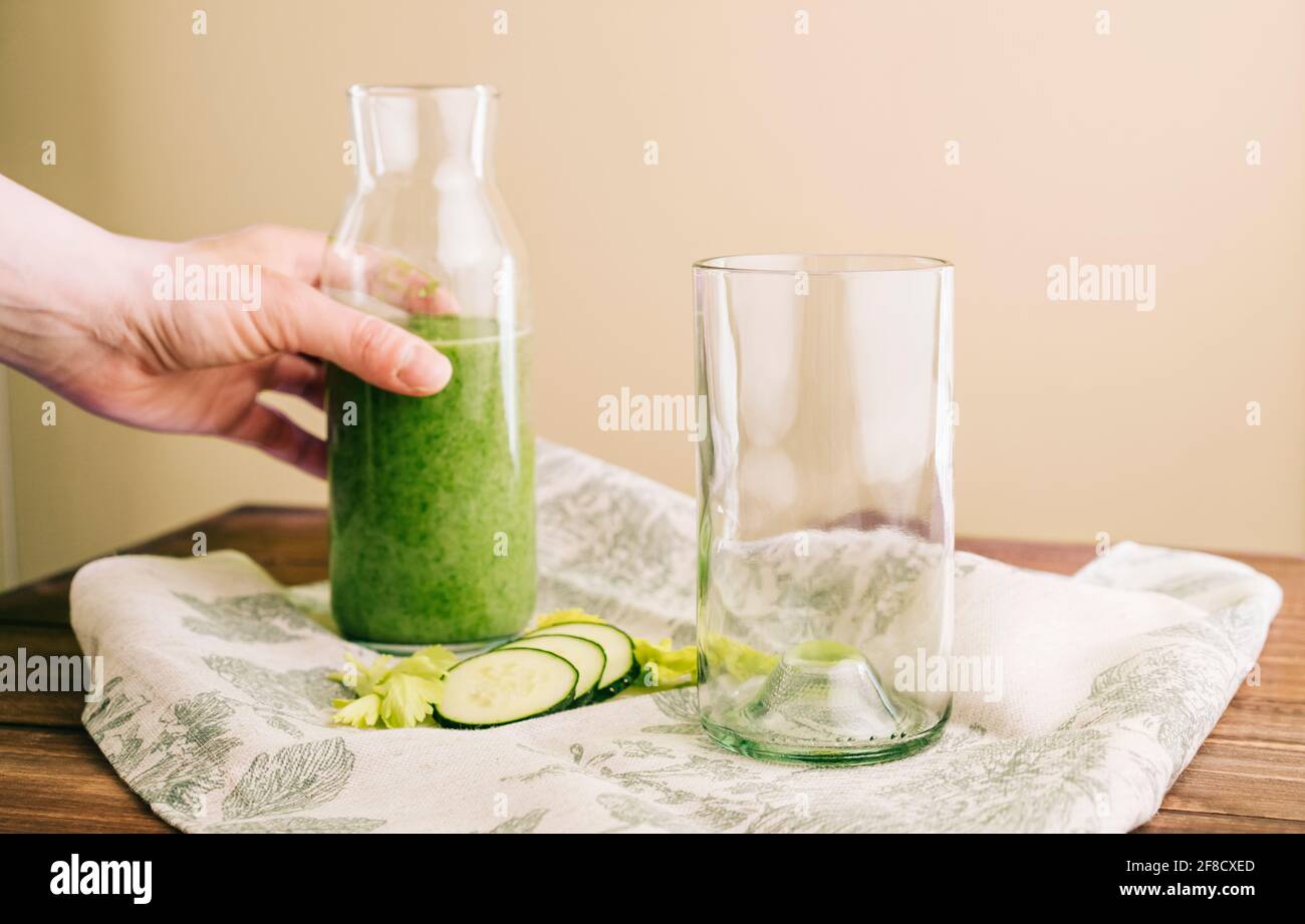 Hand hält eine Glasflasche mit grünem Smoothie und Kristall Glas auf einem grauen Tuch mit Sellerie- und Gurkenscheiben Stockfoto