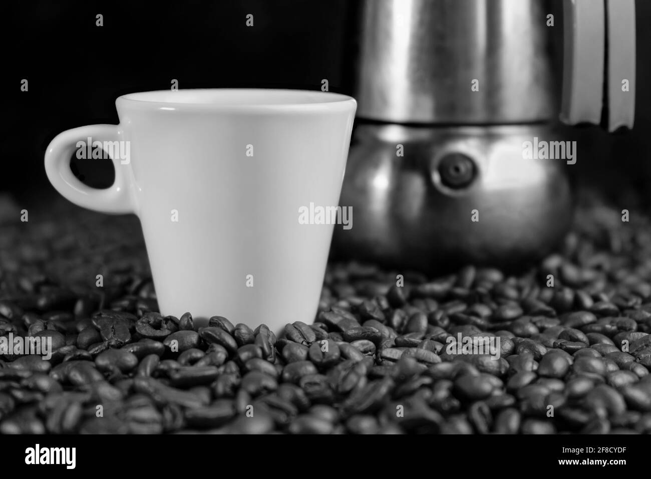 Schwarzweiß-Foto mit einer italienischen Espressomaschine, Kaffeebohnen und einer weißen Tasse auf dunklem Hintergrund. Stockfoto