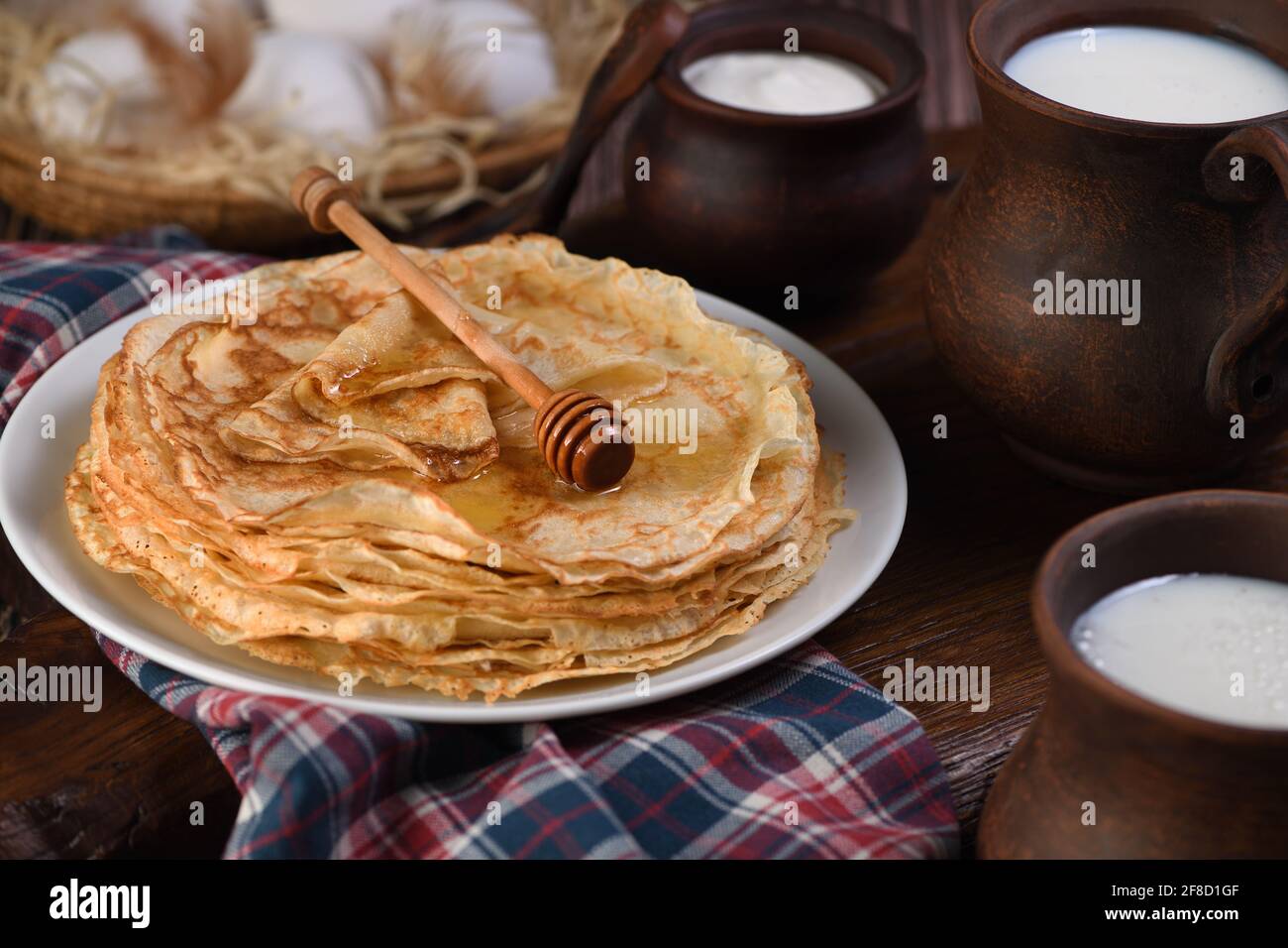 Hausgemachte dünne Pfannkuchen mit Honig, gestapelt in einem Stapel, auf  einem Holztisch mit einem Becher Milch, einem Topf mit saurer Rahm und  Eiern in einem Korb. Traditionell Slawisch Stockfotografie - Alamy