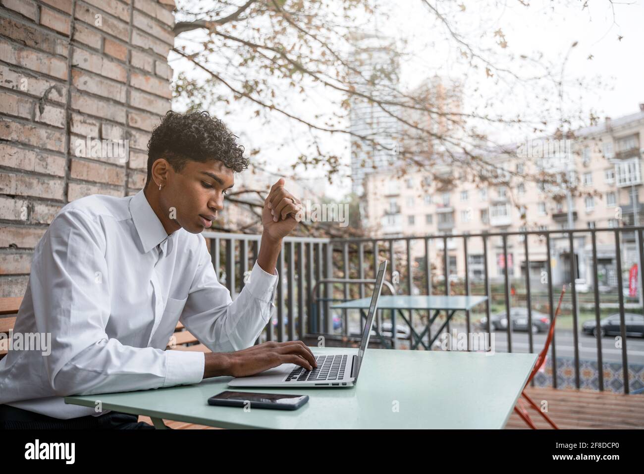 Ein schwarzer Mann sitzt auf einer Sommerterrasse oder Veranda in der Nähe eines Cafés. Der Typ arbeitet an einem Laptop. Blick auf das Frühlingsgeschäft in der Innenstadt Stockfoto