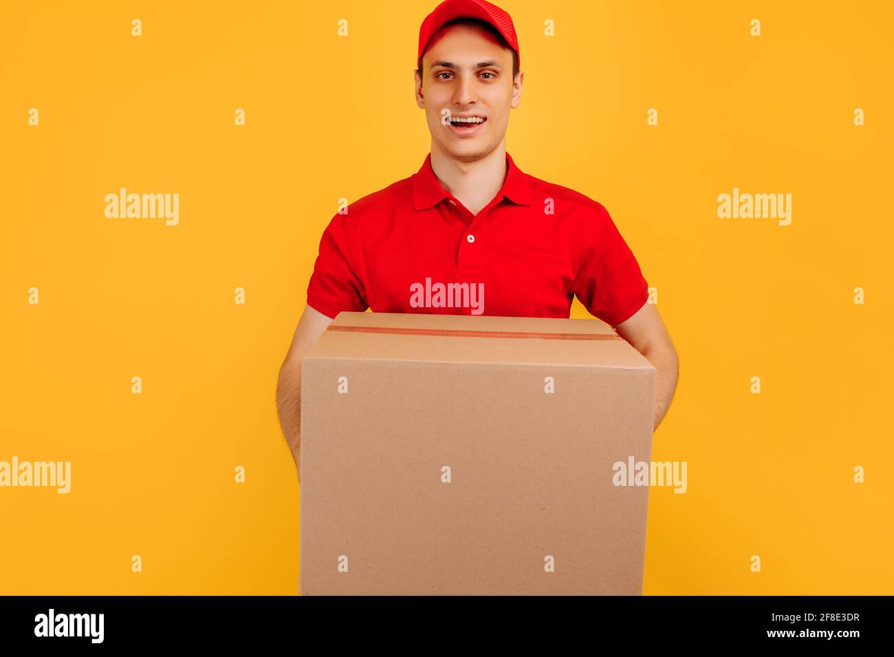 Fröhlicher, aufgeregt männlicher Kurier mit rotem T-Shirt und Mütze, der das Paket auf einem isolierten gelben Hintergrund hält, Express-Lieferung, Einkaufskonzept Stockfoto