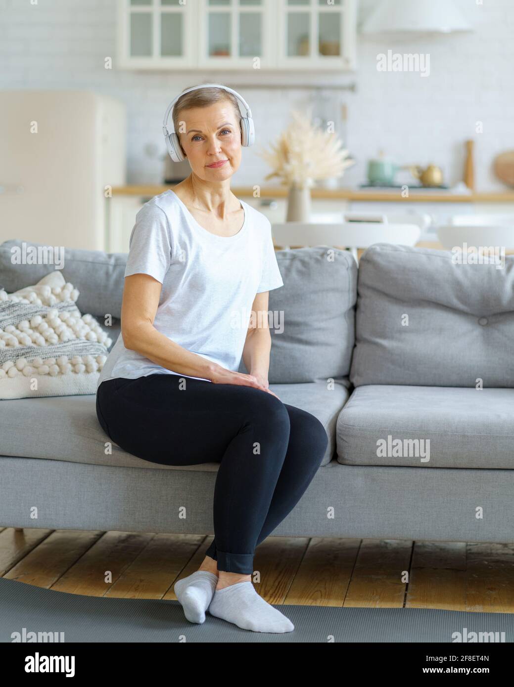 Porträt einer älteren Frau, die auf die Kamera schaut Sitzen Sie auf dem Sofa in Kopfhörern und hören Sie im Wohnzimmer Musik. Entspannen Sie sich nach dem Fitnesstraining. Emotionale und spirituelle Gesundheit. Wohlbefinden, Wellness für pensionierte Dame Stockfoto
