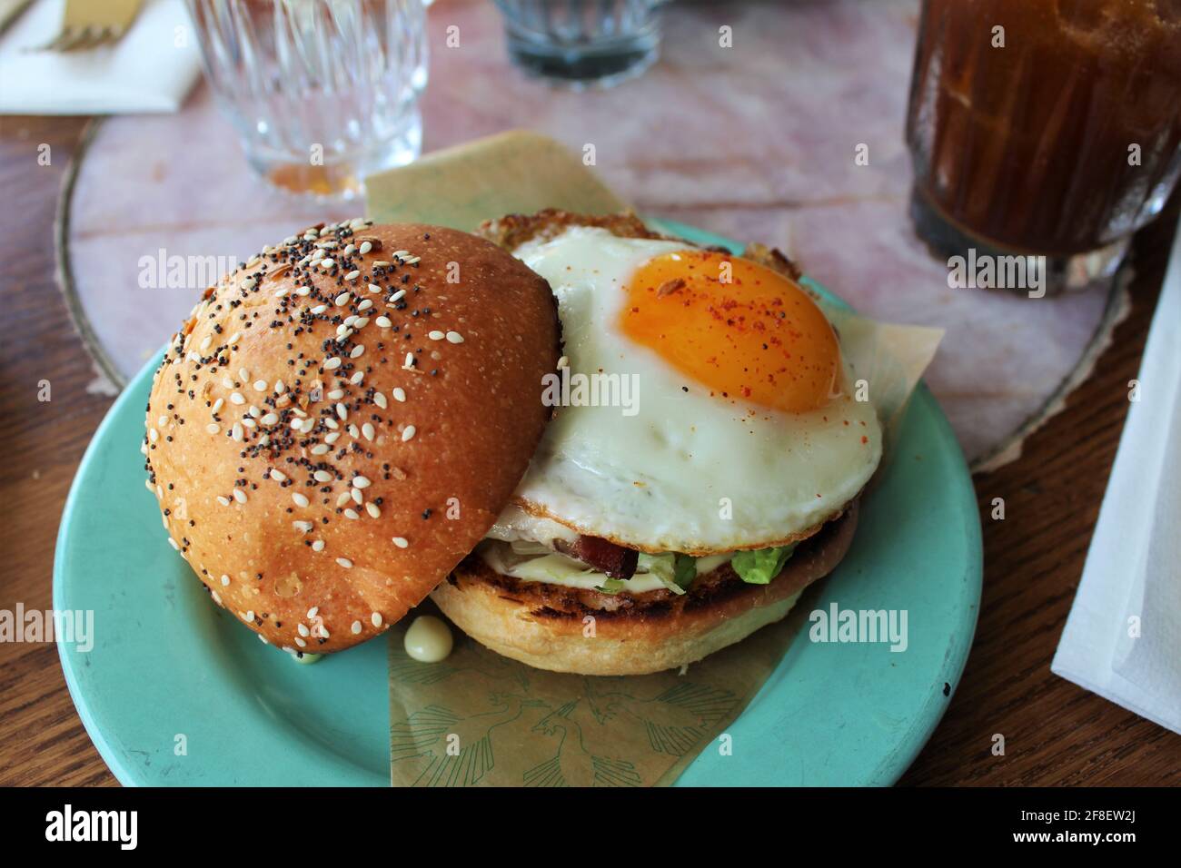 Frühstückssandwich mit einem Ei auf der Sonnenseite in einem Brioche-Brötchen mit allem Gewürz und Mohnsamen und auf einen blaugrün gefärbten Teller gelegt. Brunch Stockfoto