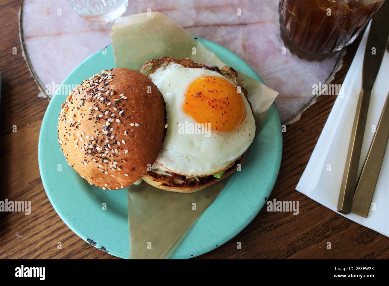 Heißer, leckerer Burger mit Ei auf der sonnigen Seite auf einem Brioche-Brötchen mit Mohnsamen und Gewürzen auf einem blaugrün gefärbten Teller. Stockfoto