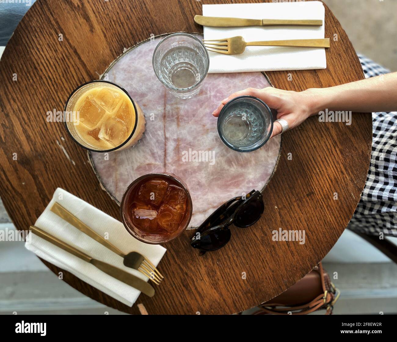 Tasse Eiskaffee mit Macadamia-Milch und Kräutertee-Eistee auf einem Holztisch mit Gold Besteck und die Hand einer Frau, die eine Cu Stockfoto