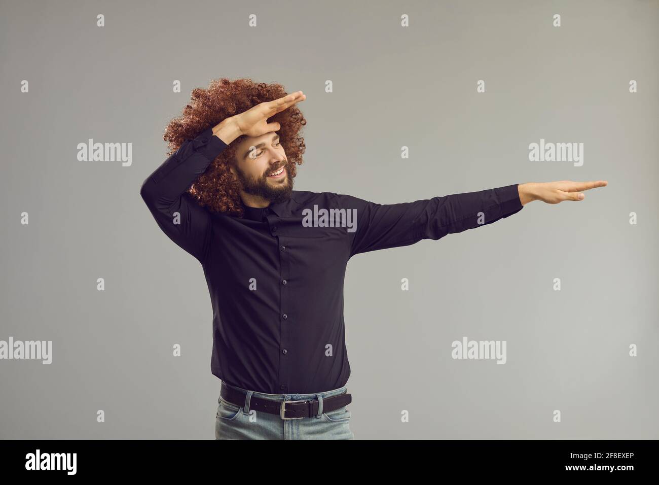 Lustiger Kerl in großer lockiger Perücke, der tuckende Tanzbewegung macht Im Studio mit grauem Hintergrund Stockfoto