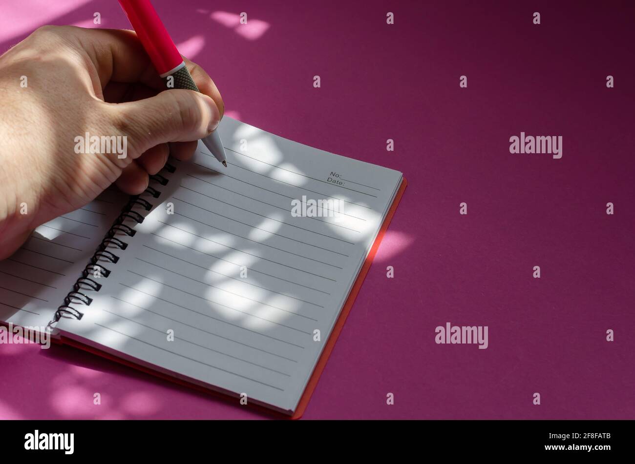 Licht- und Schattenkomposition der linken Hand in einem Notizbuch auf einem rosa Hintergrund. Ein Mann mittleren Alters macht Notizen mit einem rosa Stift. Business, dai Stockfoto