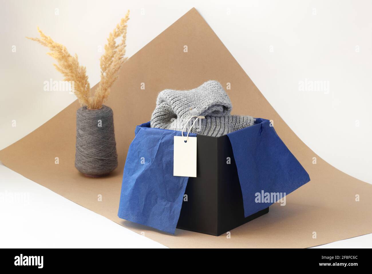 Weißes, leeres Papier-Logo auf einem grauen Strickschal In einer schwarzen Schachtel und blauem Seidenpapier und getrocknet pampas Gras in einer Vase aus Spulengarn auf beigefarbenem Papier Stockfoto