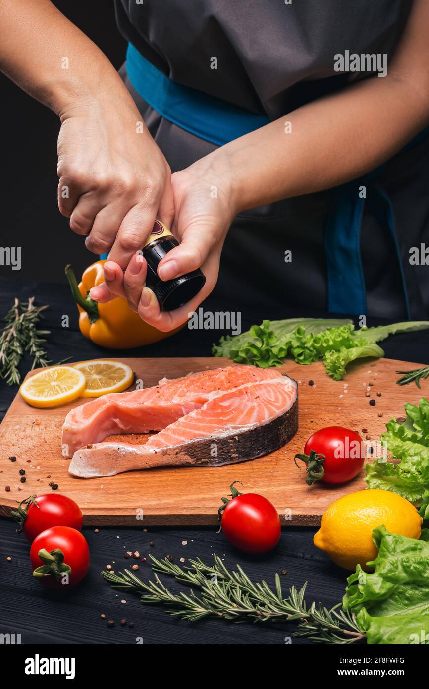 Kochen von Lachs. Weibliche Hände streuen Lachsfischsteak mit Gewürzen auf ein schönes Holzbrett. Zutaten Zitrone, Rosmarin, Pfefferkörner, Tomaten, p Stockfoto