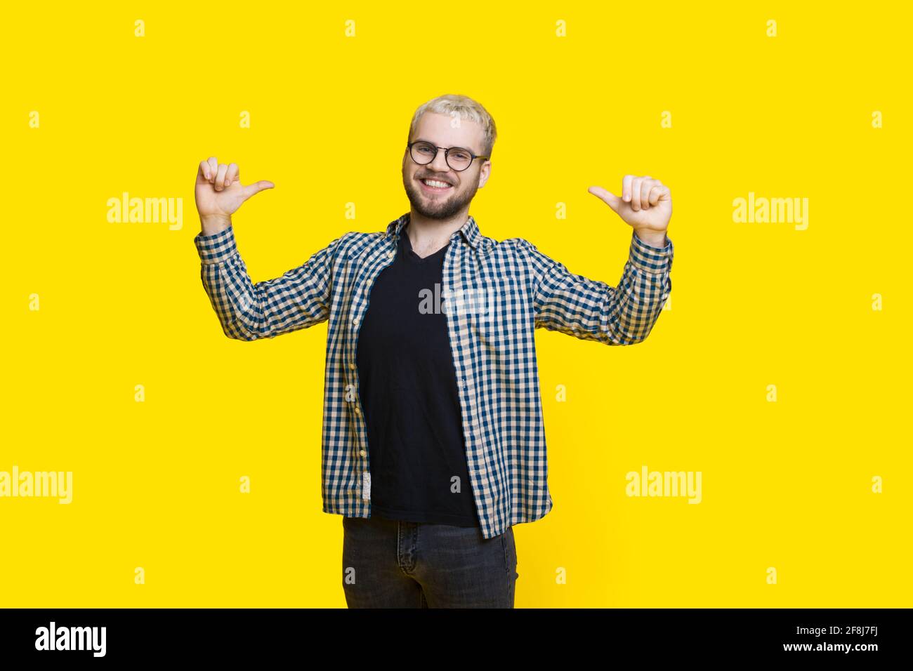 Selbststolzer Mann gestikuliert sich lächelnd auf einem Gelbe Studiowand durch eine Brille mit Bart Stockfoto