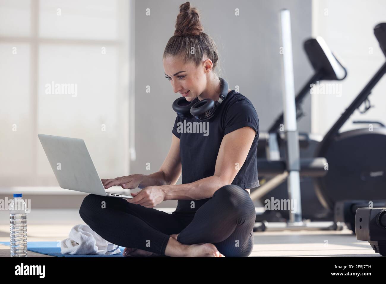 Eine sportliche Frau in Sportbekleidung sitzt auf dem Boden Mit Hanteln und einem Proteinshake oder einer Flasche Wasser und wird mit einem Laptop zu Hause in der livi Stockfoto