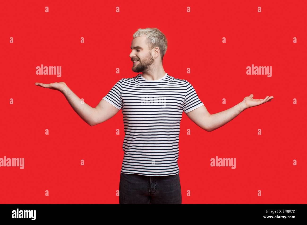 Bärtiger blonder Mann vergleicht die Dinge auf seinen Handflächen, die posieren Und lächelnd in einer roten Studiowand Stockfoto