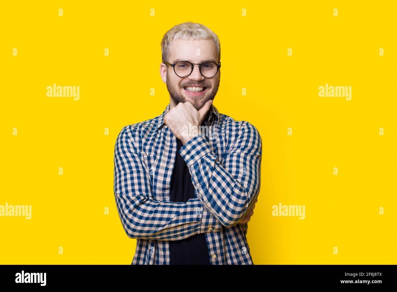 Der kaukasische Mann berührt sein bärtiges Kinn und lächelt der Kamera zu An einer gelben Studiowand mit Brille und Hemd Stockfoto