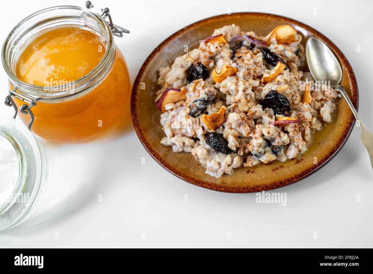 Haferbrei mit Apfelstücken, getrockneten Pflaumen und Honig in Teller mit Löffel und Glasschüssel mit Honig auf weißem Hintergrund. Gesundes, süßes Frühstück. Stockfoto