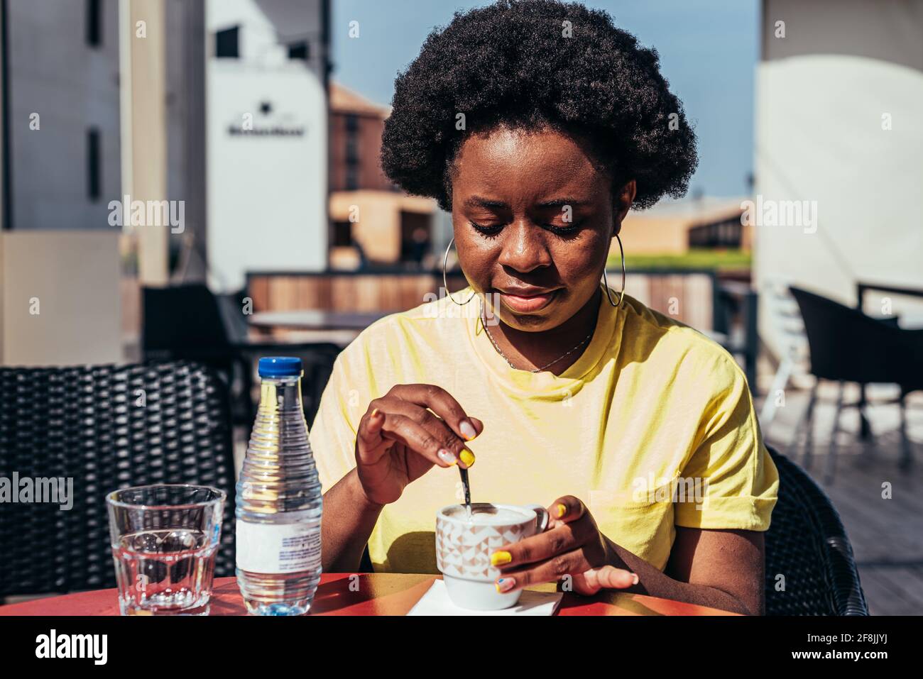 Porträt eines schwarzen Mädchens mit Afro-Haaren und Reifringen, das Kaffee und eine Flasche Wasser auf einer Barterrasse trinkt. Stockfoto