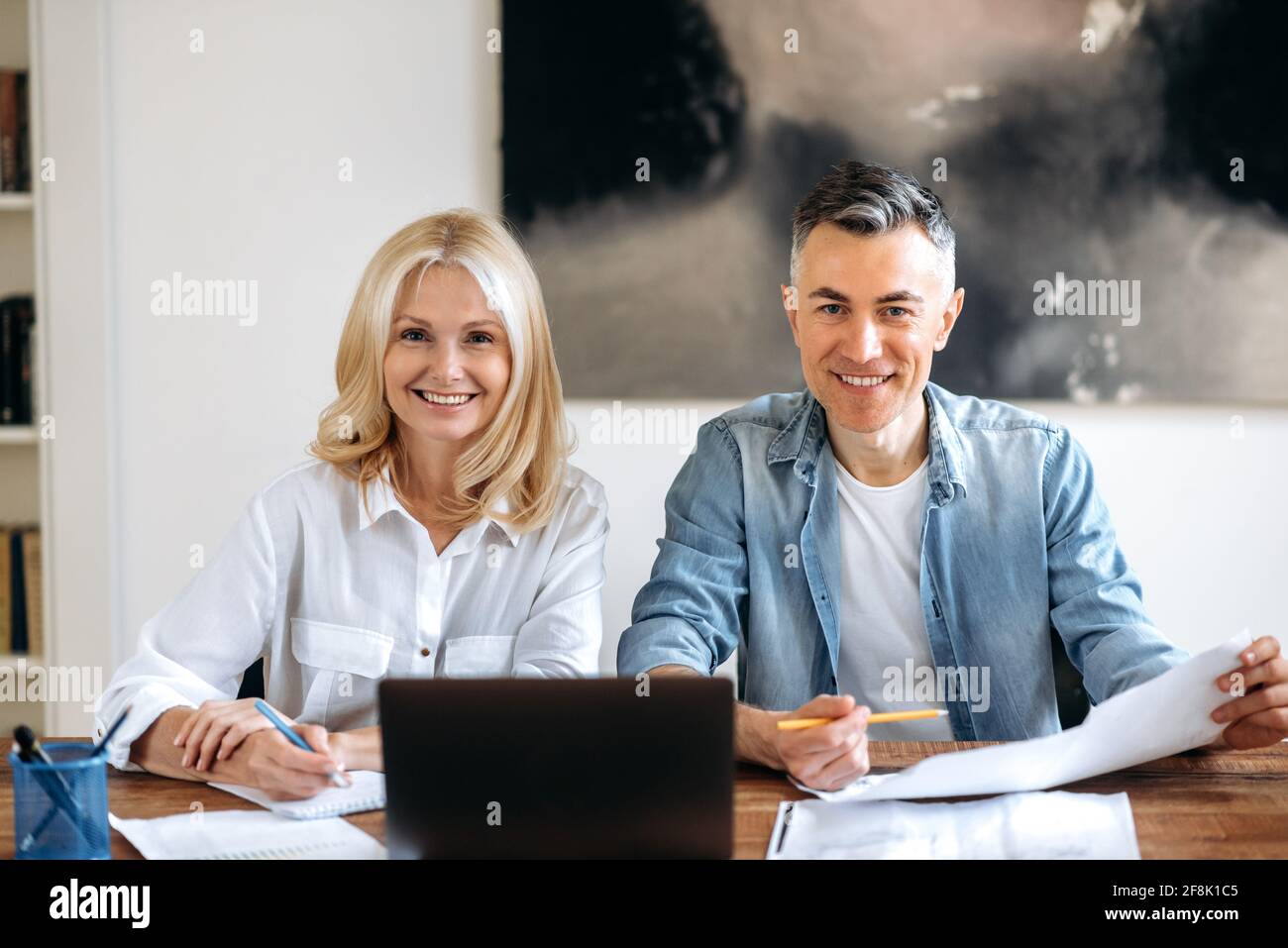 Intelligenter Geschäftsmann und Geschäftsfrau. Zwei erfolgreiche Partner sitzen am Arbeitsplatz, arbeiten an einem neuen Projekt, nutzen einen Laptop, schauen sich Dokumente an, blicken freundlich lächelnd auf die Kamera Stockfoto
