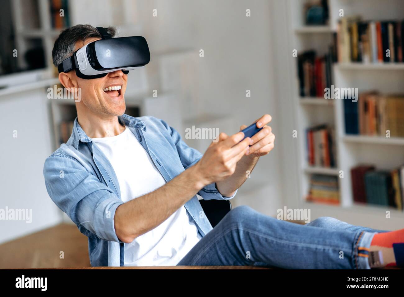 Aufgeregt mittleren Alters kaukasischen Mann sitzt in am Schreibtisch und gestikulieren Hände beim Testen der neuen App über VR-Gerät. Fröhlicher reifer Mann mit VR-Brille und hält Smartphone, Virtual-Reality-Konzept Stockfoto